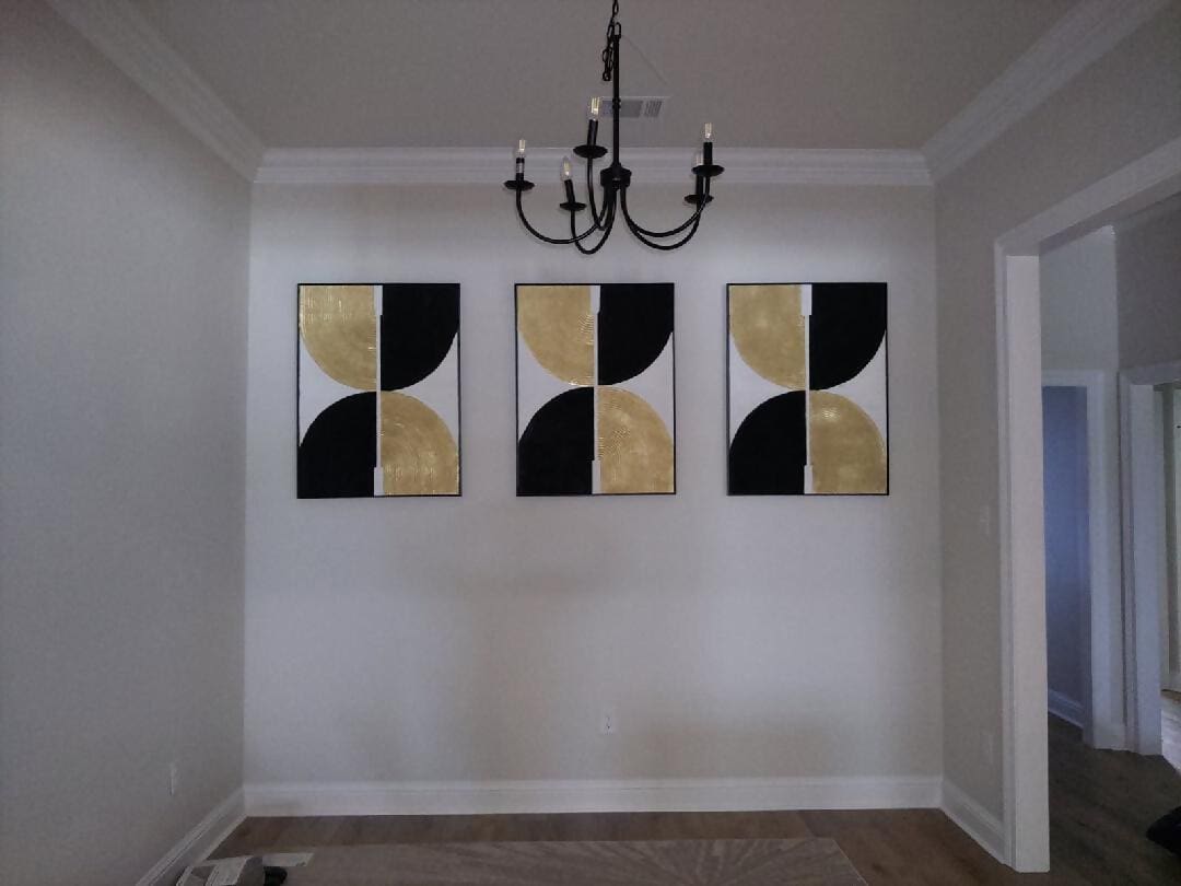Three abstract black and gold paintings hang on a white wall under a chandelier.