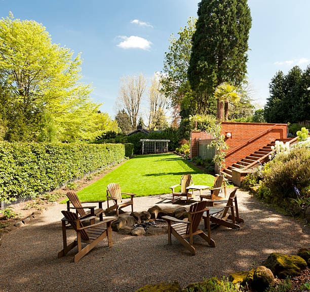 A sunny backyard with green lawn and wooden chairs around a fire pit.