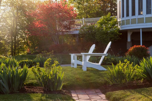 Two white Adirondack chairs on a sunny garden lawn with greenery and a brick path.