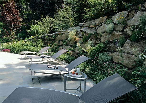 Lounge chairs with side tables set beside a stone wall in a lush garden.