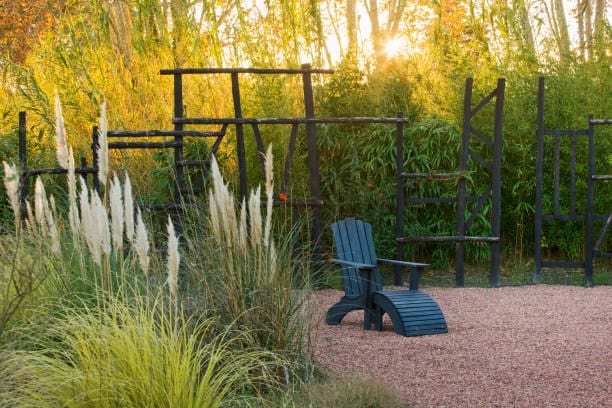 Two blue Adirondack chairs on a gravel path amid tall grasses at sunset.